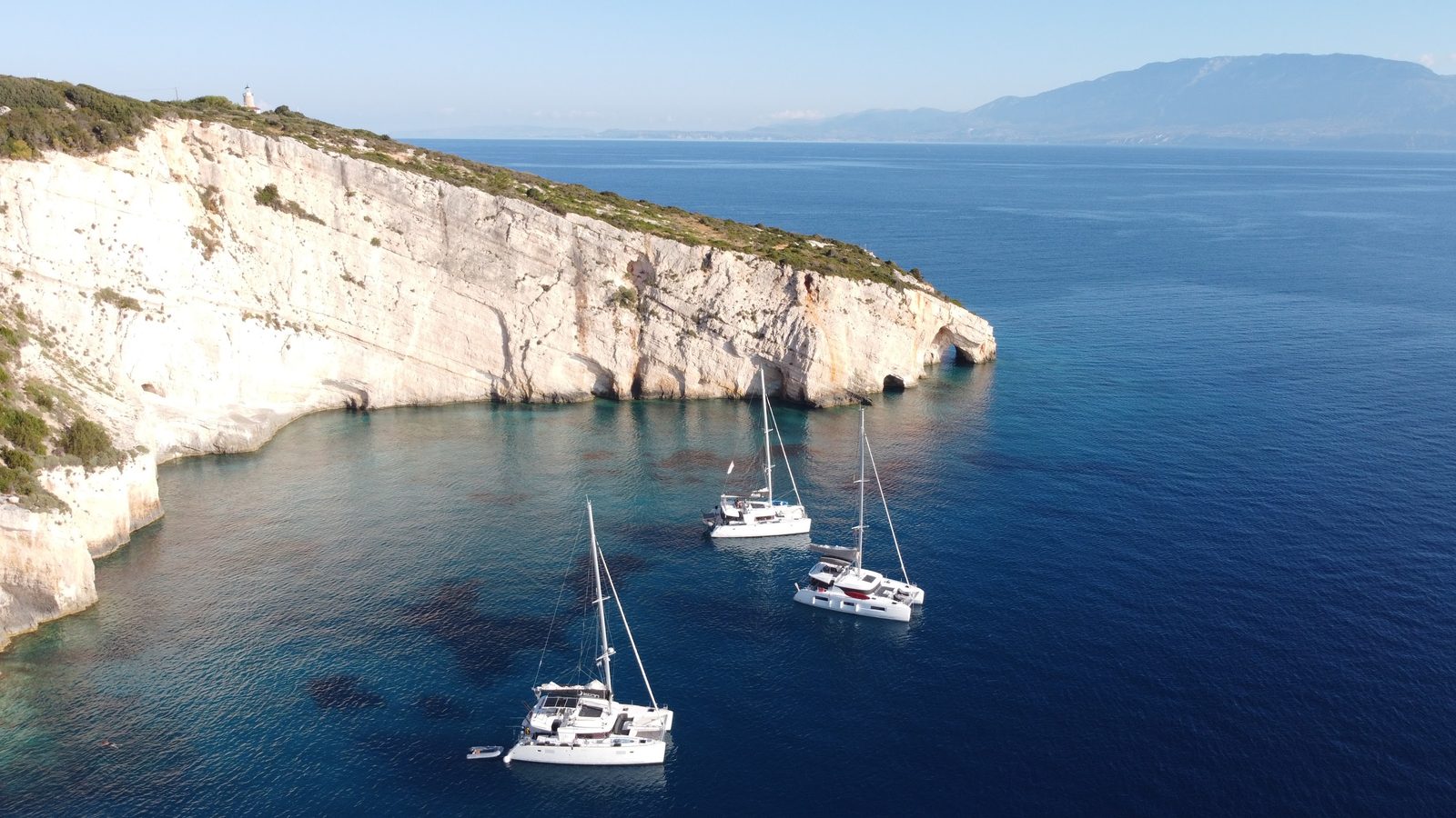 Three catamarans at anchor under the white cliffs of the Blue Caves, the Skinari lighthouse above