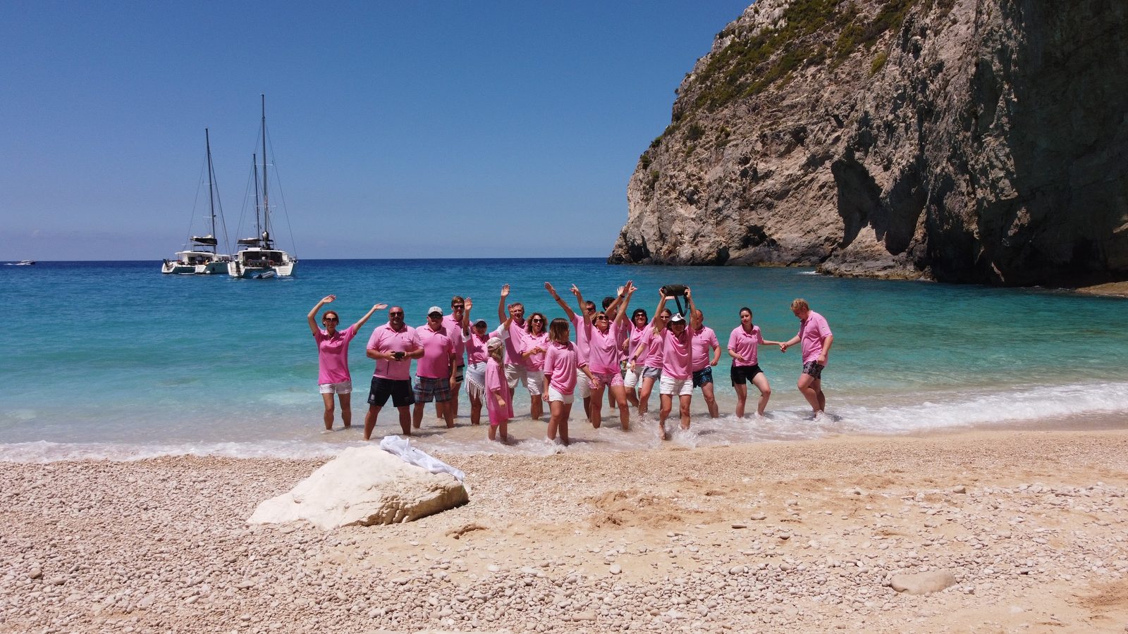 A group in matching pink T-shirts on a pebble beach, their two catamarans anchored behind