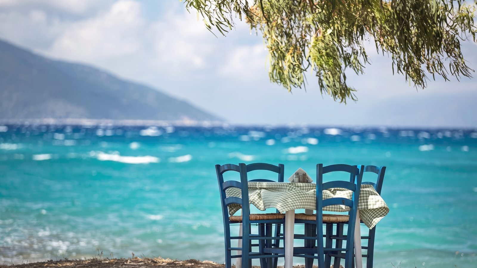 A taverna table under a tree on the shore at Sami, the bay and mountains behind