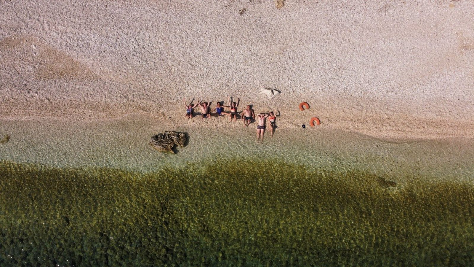 The group lying on the pebbles at the water's edge, seen from the drone