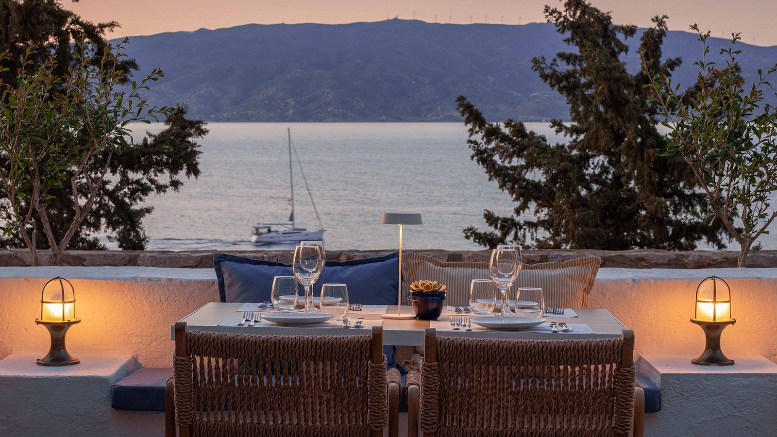 A terrace table set for dinner at dusk, a yacht at anchor beyond the pines