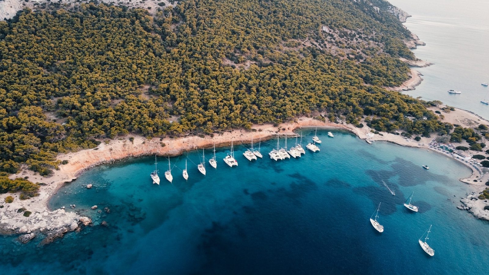 Yachts anchored off Moni islet from the air, pine forest down to a pale cove